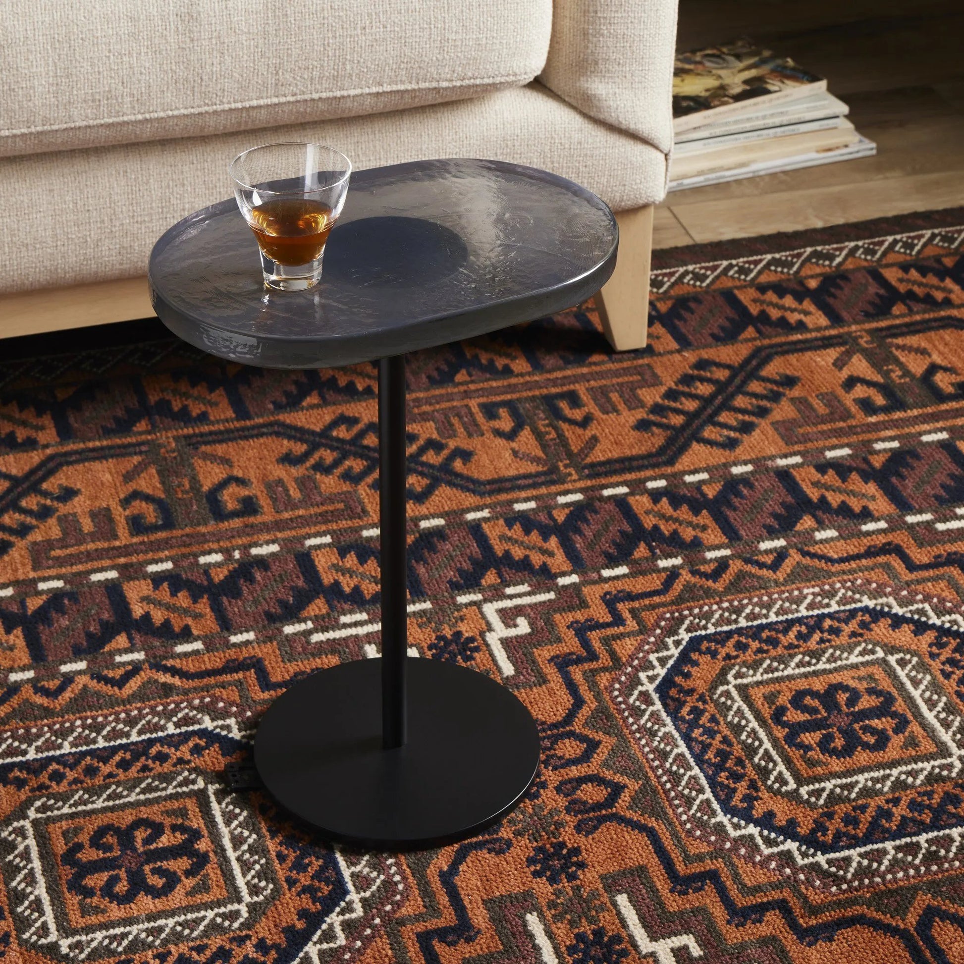 A slim-profile end table pairs a simple iron base with an oval tabletop of hand-cast recycled glass in a soft, smoky blue Amethyst Home provides interior design, new home construction design consulting, vintage area rugs, and lighting in the Winter Garden metro area.